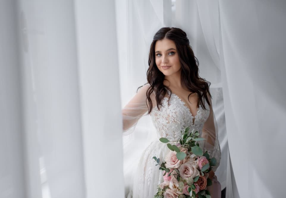 Smiling bride holding a bouquet near a window with sheer curtains
